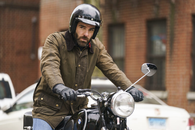 Zachary Quinto, a bearded man is riding a motorcycle, wearing a black helmet and brown jacket with a corduroy collar. His expression is serious, and he has black leather gloves on. The motorcycle is vintage-style, with a large round headlight and a visible BMW logo. The background shows a brick building and a parked white car, giving an urban setting to the scene.