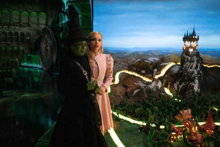 In a scene from Wicked, two characters stand together, one dressed in black with green skin and a pointed hat, and the other in soft pink attire with blonde hair. They overlook a glowing, fantasy-inspired landscape with winding paths, small houses, and a castle on a mountain.