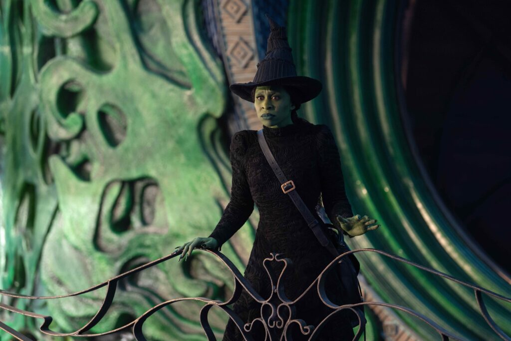 A scene from Wicked shows a close-up of a character with green skin and a pointed black hat, dressed in a textured black outfit. They are standing on an ornate, curved balcony in front of an intricately designed green background.