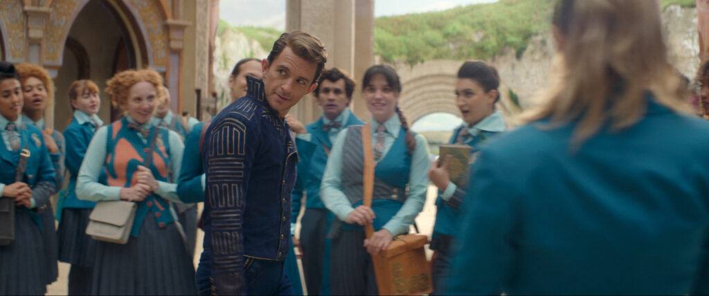 A scene from Wicked shows a group of students in teal uniforms gathers outdoors near an arched building. A man in a navy blue jacket with gold patterns stands in the foreground, looking back with a confident expression, while the students watch him with interest.