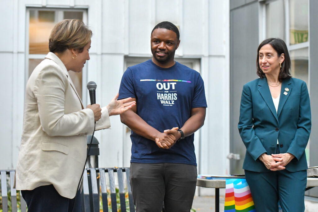 From left, Massachusetts Governor Maura Healey speaks into a microphone while gesturing towards State Rep. Andre Carroll, who stands beside her with hands clasped, wearing a navy T-shirt that reads “OUT for HARRIS WALZ.” On the right, City Councilmember Rue Landau looks on, dressed in a teal suit. The group appears to be at an outdoor event, with a bench and a table draped with a rainbow flag in the background.