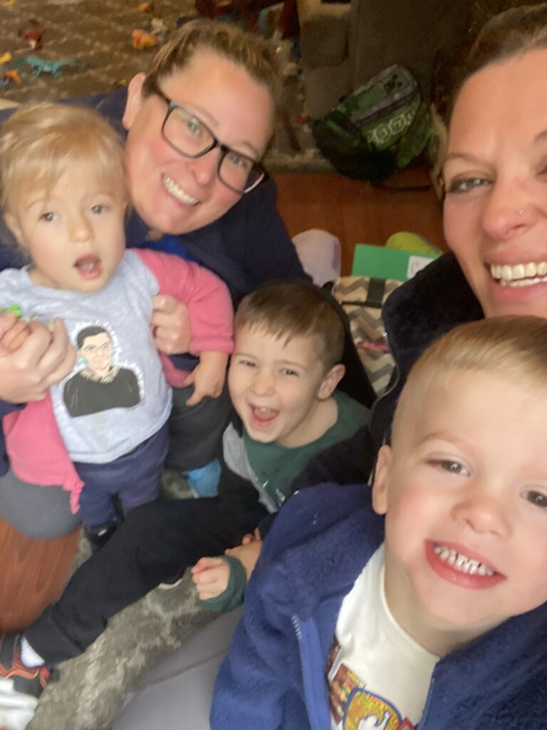 A colorful image of two women and three young children sitting together indoors. One woman, wearing glasses, holds a toddler, while the other leans in, smiling. The children are laughing and appear happy, surrounded by toys on the floor.