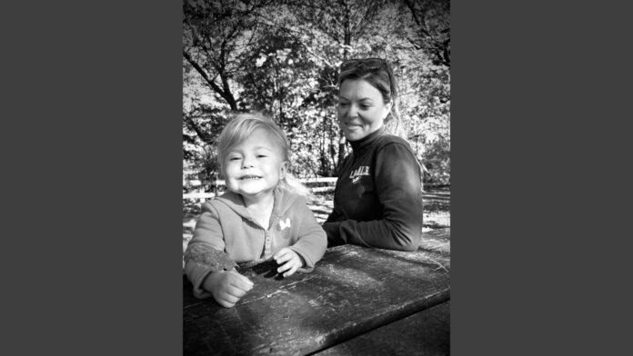 A black-and-white photo of a young child and a woman outdoors at a picnic table. The child is smiling brightly and holding a leaf, while the woman sits beside them, looking toward the child. Trees and sunlight are visible in the background.