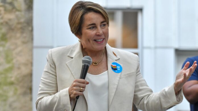 Massachusetts Governor Maura Healey speaks while holding a microphone. She is wearing a beige blazer over a white top, with a blue circular pin on her lapel that reads “OUT” in white text, accompanied by a rainbow-colored stripe design. Her facial expression is friendly, and she gestures with one hand. The background is softly blurred, highlighting her as the focal point.