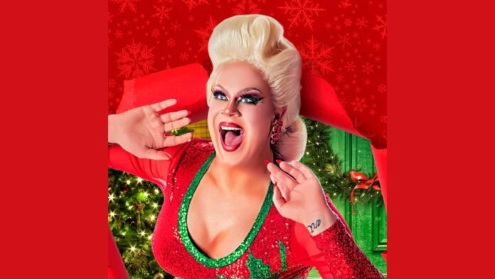 Nina West in festive holiday attire, wearing a sparkly red dress with green trim, styled in a retro look with bold makeup and platinum blonde hair. She has a wide smile and open hands beside her face, with a background featuring Christmas trees, green and red decorations, and a red snowflake-patterned backdrop.