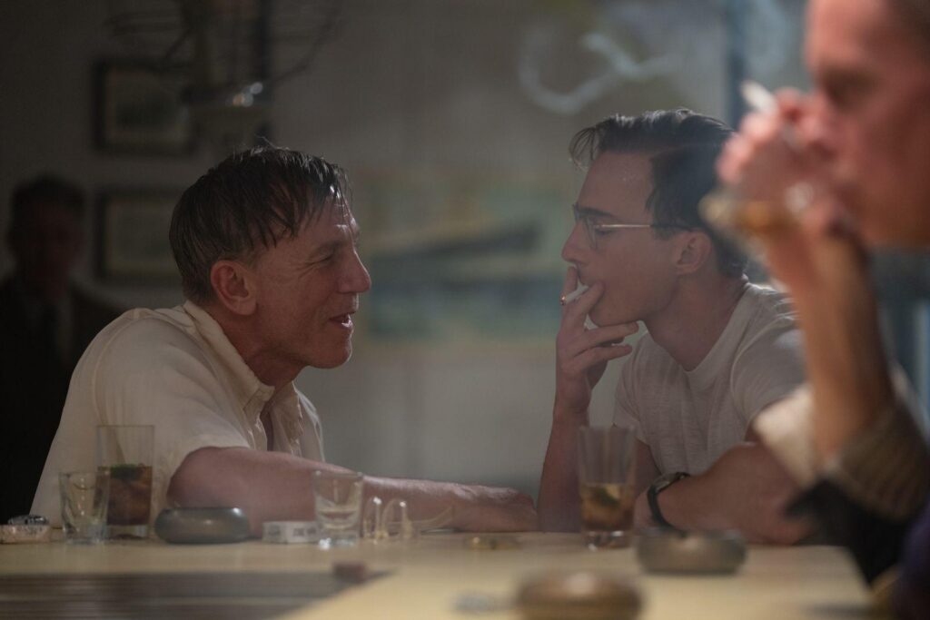 Daniel Craig and Drew Starkey share an intense moment in Queer, sitting across a bar table amidst a haze of smoke and empty glasses. Their expressions suggest a mix of longing and tension, highlighting the complex dynamic between their characters.