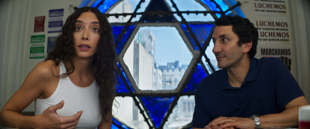 Penélope Guerrero, dressed in a white tank top, gestures as she speaks to Juan Minujín, who listens attentively, sitting beside her. The background features a large blue stained-glass Star of David window and posters with Spanish text about rights and pride. This is a scene from Transmitzvah.