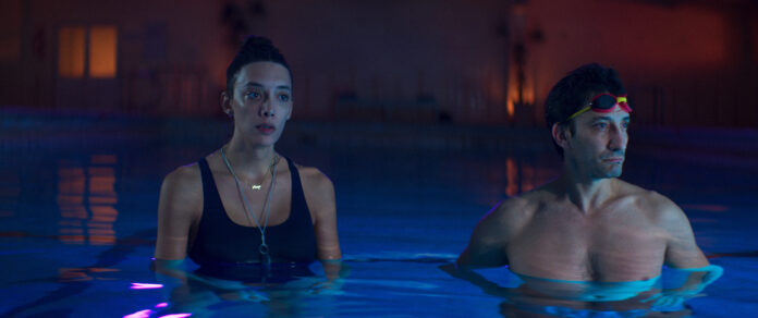 Penélope Guerrero and Juan Minujín stand waist-deep in a dimly lit swimming pool, their expressions contemplative. Guerrero wears a black swimsuit with layered necklaces, while Minujín has goggles perched on his forehead. The pool reflects soft blue and pink lighting. This is a scene from Transmitzvah.