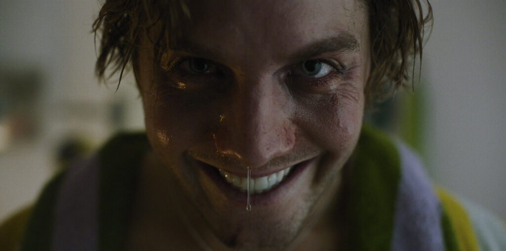 A close-up image of Lukas Gage with messy, slightly wet hair and a wide, intense smile. His face is glistening, likely from sweat, and a small droplet hangs from his nose. His eyes are wide and piercing, giving him a somewhat unhinged or mischievous expression. The background is blurred, drawing focus to his face, which appears tired or worn, with visible wrinkles and dark shadows around his eyes. A scene from Smile 2.