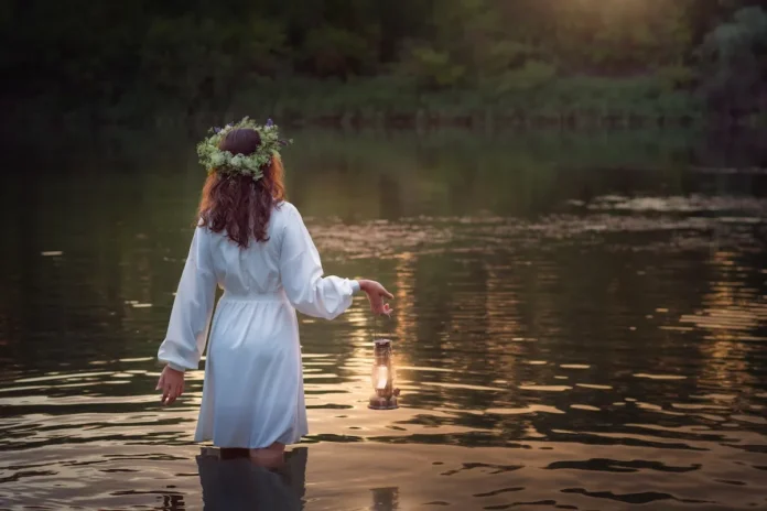 The image shows a person standing in shallow water, facing away from the camera. They are wearing a white dress and a floral crown, and they hold a lit lantern close to the water. The scene is softly lit by warm, natural light, creating a serene, almost mystical atmosphere, with trees and greenery in the background.