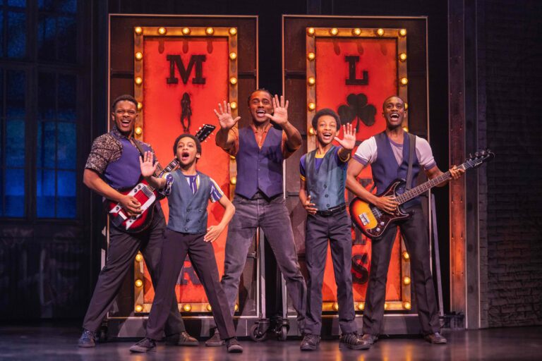 The cast of the "MJ" First National Tour portrays The Jackson 5, standing in a lively pose on stage. The group includes five performers wearing coordinated outfits with vests, smiling and gesturing energetically. Two members hold guitars, while a vibrant backdrop with illuminated signs reading "M" and "L" adds a dynamic atmosphere.