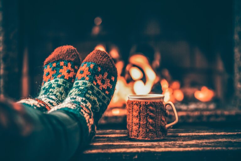 Feet in woollen socks by the Christmas fireplace. Woman relaxes by warm fire with a cup of hot drink and warming up her feet in woollen socks. Close up on feet. Winter and Christmas holidays concept.