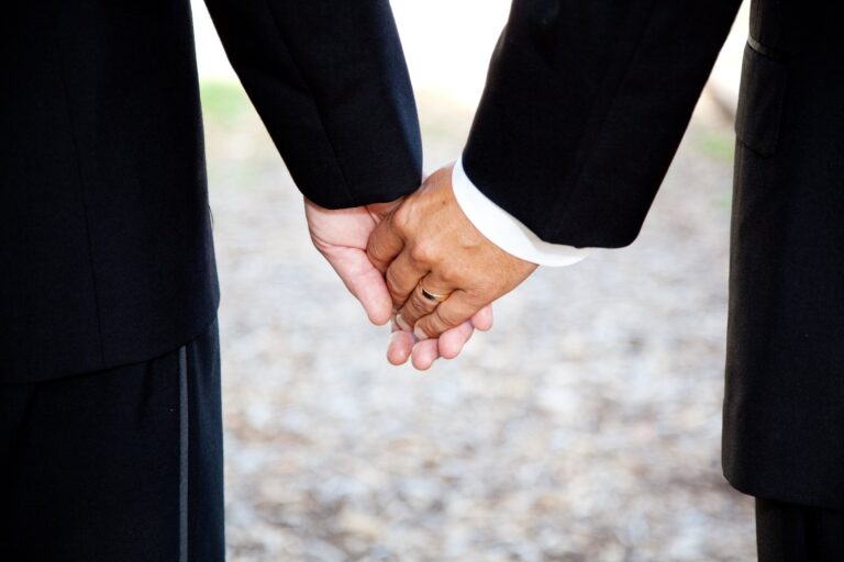 Closeup of a gay couple holding hands, wearing a wedding ring. Couple is a hispanic man and a caucasian man.