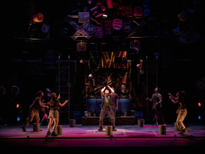 A vibrant stage performance from STOMP, featuring six performers energetically interacting with everyday objects as percussion instruments. The set is adorned with industrial and recycled items like road signs, barrels, and scrap metal, creating a visually striking background. The performers, illuminated by colorful stage lighting, are actively engaged, with one in the center tossing a tin can mid-air while others reach towards it. The scene captures the dynamic and rhythmic energy characteristic of STOMP.