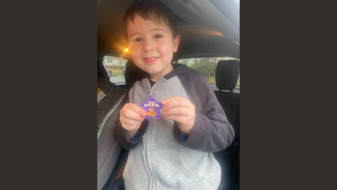 A young boy smiling while holding a purple star-shaped keychain that says "UMPS R.O.A.R. - Spawns Pride" inside a car, with a car seat visible in the background.