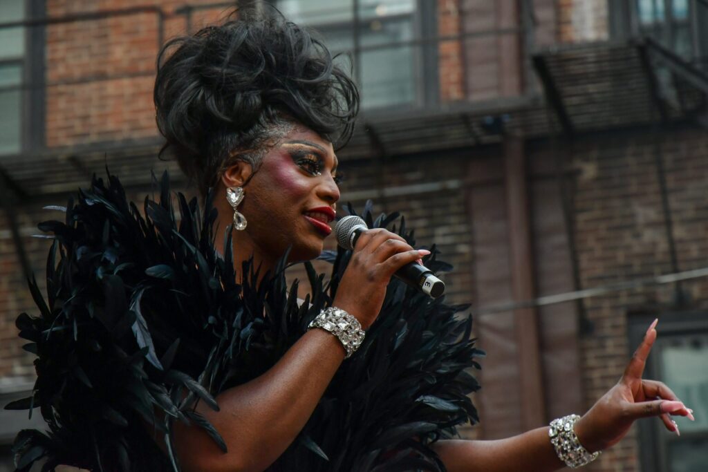 Drag performer Sapphira Cristál, wearing a dramatic black feathered outfit and sparkling jewelry, sings passionately into a microphone against a backdrop of urban brick buildings.