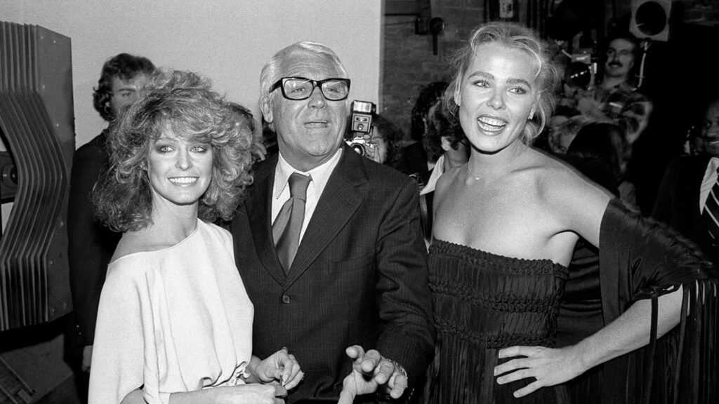 A black-and-white image from the documentary Studio One Forever featuring three individuals at a glamorous event. On the left is a woman with voluminous, wavy hair and a bright smile, wearing an elegant off-shoulder top. In the center, an older man in glasses and a suit smiles warmly, and on the right, a woman in a strapless dress with a fringed shawl beams with joy. The lively background captures other attendees and photographers, adding to the celebratory atmosphere.