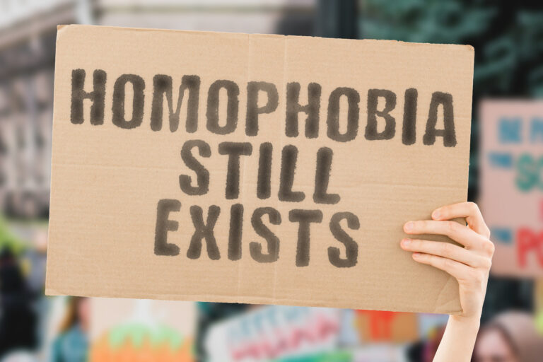 The phrase " Homophobia still exists " on a banner in men's hand. Human holds a cardboard with an inscription. Humanity. Difference. Tolerant. Negative. Civil rights. Negativity. Conflict