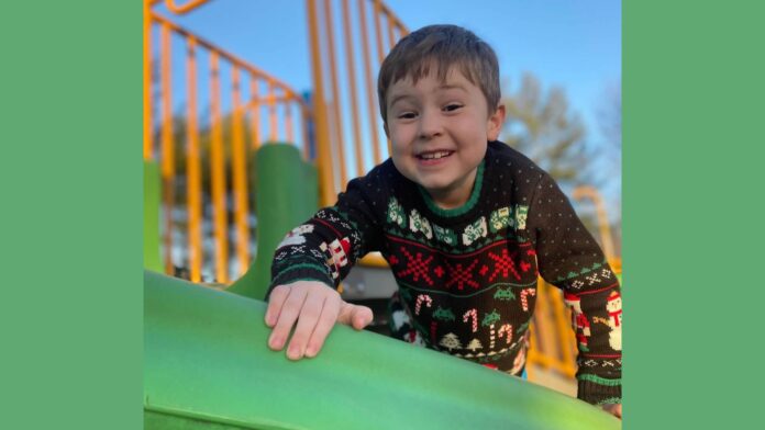 A young boy with short brown hair and a wide smile is climbing on a green play structure at a playground. He is wearing a festive black sweater with Christmas-themed patterns, including snowmen, candy canes, and trees. His hand rests on the edge of the structure as he looks at the camera. The background features yellow playground bars and a clear blue sky.