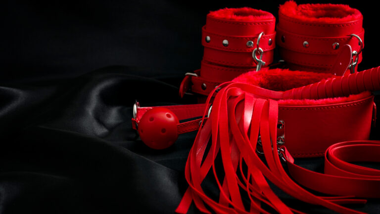 Bondage, kinky adult sex games, kink and BDSM lifestyle concept with a pair of red leather handcuffs, flogger, ball gag and a coller with a leash attached on black silk with copy space