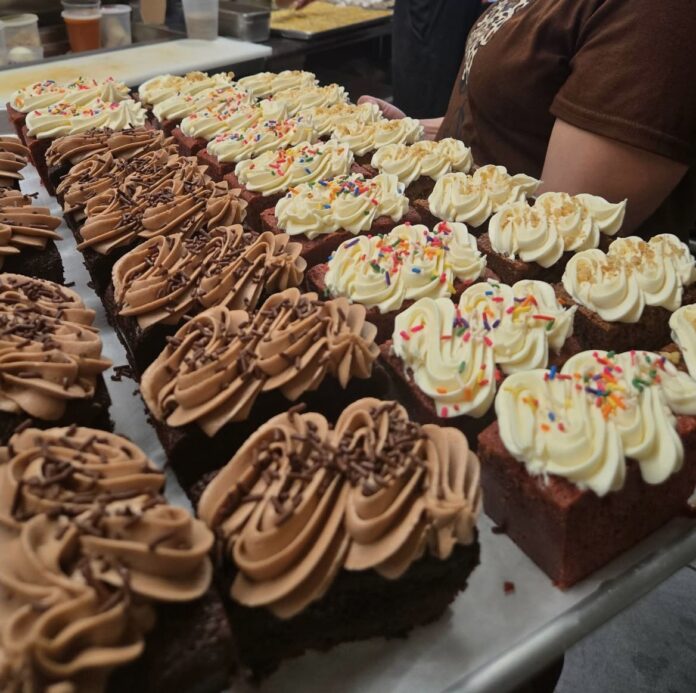 Darnel's Cakes presents a tray of freshly baked square-shaped cupcakes is displayed, topped with swirls of frosting. The front row features chocolate cupcakes with light brown chocolate frosting and chocolate sprinkles. The back rows have red velvet cupcakes with creamy white frosting, some adorned with colorful sprinkles and others with crumbled toppings. A person wearing a brown shirt is partially visible behind the tray in a commercial kitchen setting.